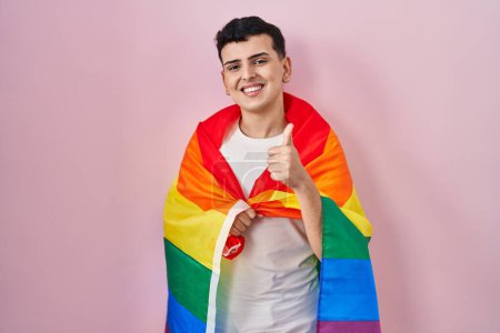 Photo for Non binary person holding rainbow lgbtq flag smiling happy and positive, thumb up doing excellent and approval sign - Royalty Free Image