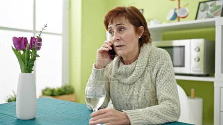 Photo for Mature hispanic woman speaking on the phone sitting on the table drinking a glass of white wine at dinning room - Royalty Free Image