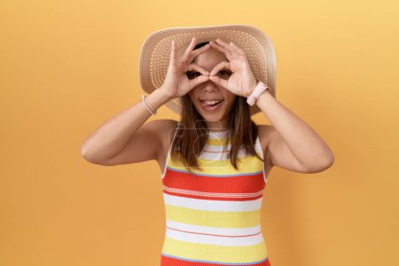Photo for Middle age chinese woman wearing summer hat over yellow background doing ok gesture like binoculars sticking tongue out, eyes looking through fingers. crazy expression. - Royalty Free Image