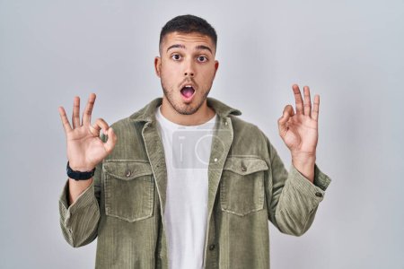 Téléchargez les photos : Young hispanic man standing over isolated background looking surprised and shocked doing ok approval symbol with fingers. crazy expression - en image libre de droit