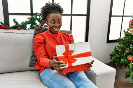 Foto de African american woman unpacking christmas gift sitting on sofa at home - Imagen libre de derechos