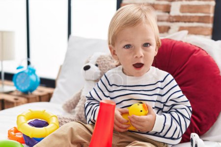 Téléchargez les photos : Adorable garçon caucasien jouant avec cerceaux jeu tenant jouet de canard assis sur le lit dans la chambre - en image libre de droit
