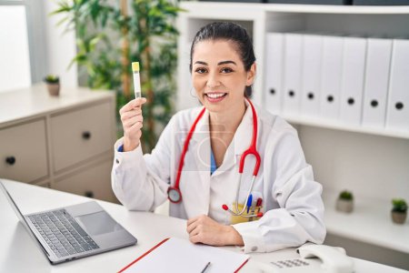 Photo for Young doctor woman holding blood sample looking positive and happy standing and smiling with a confident smile showing teeth - Royalty Free Image