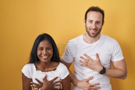 Téléchargez les photos : Couple Interracial debout sur fond jaune souriant et riant fort à haute voix parce que drôle blague folle avec les mains sur le corps. - en image libre de droit