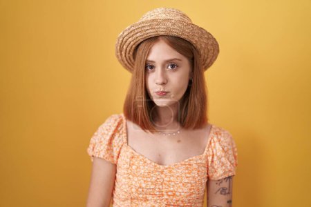 Téléchargez les photos : Jeune femme rousse debout sur fond jaune portant un chapeau d'été gonflant les joues avec un visage drôle. bouche gonflée d'air, expression folle. - en image libre de droit