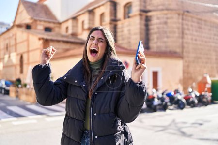 Téléchargez les photos : Jeune belle femme hispanique utilisant un smartphone avec expression gagnante dans la rue - en image libre de droit