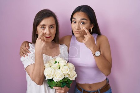 Téléchargez les photos : Mère et fille hispaniques tenant un bouquet de fleurs blanches pointant vers l'œil vous regardant geste, expression suspecte - en image libre de droit