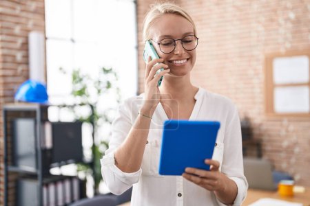 Téléchargez les photos : Jeune femme blonde travailleur d'affaires en utilisant touchpad parler sur smartphone au bureau - en image libre de droit