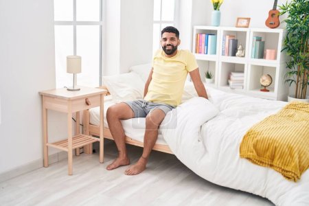 Téléchargez les photos : Jeune homme arabe souriant confiant assis sur le lit dans la chambre - en image libre de droit