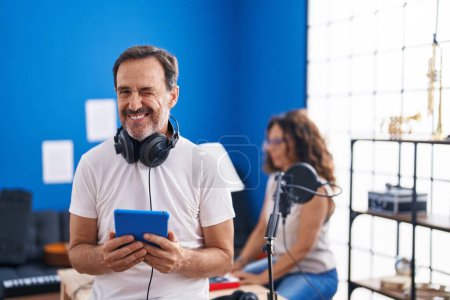 Téléchargez les photos : Moyen Age hispanique homme au studio de musique en utilisant tablette clin d'oeil en regardant la caméra avec expression sexy, gai et heureux visage. - en image libre de droit