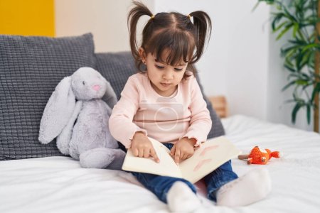 Photo for Adorable hispanic girl reading book sitting on bed at bedroom - Royalty Free Image