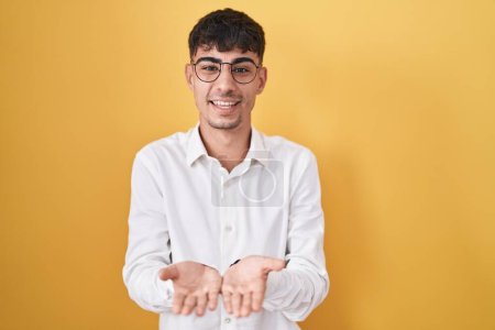 Foto de Joven hombre hispano de pie sobre fondo amarillo sonriendo con las palmas de las manos juntas recibiendo o dando gesto. retención y protección - Imagen libre de derechos