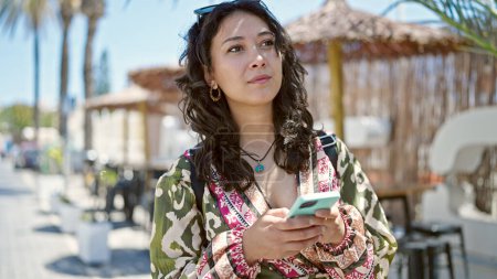Foto de Mujer hispana hermosa joven usando teléfono inteligente con expresión seria en la terraza de la cafetería - Imagen libre de derechos