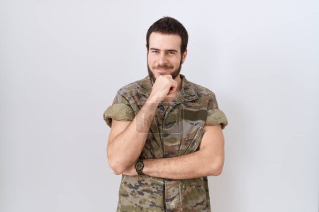 Téléchargez les photos : Jeune homme hispanique portant un uniforme de camouflage de l'armée regardant avec confiance la caméra souriant avec les bras croisés et la main levée sur le menton. penser positivement. - en image libre de droit