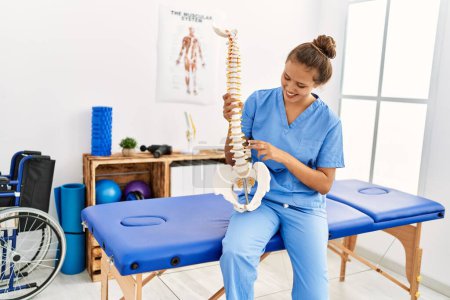 Foto de Joven bella fisioterapeuta hispana sonriendo confiada sosteniendo modelo anatómico de columna vertebral en clínica de rehabilitación - Imagen libre de derechos