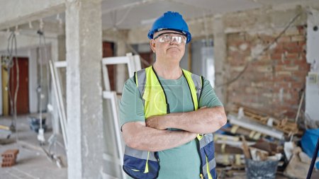 Foto de Constructor hombre de pelo gris de mediana edad de pie con expresión relajada y brazos cruzados gesto en el sitio de construcción - Imagen libre de derechos