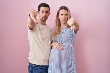 Photo for Young couple expecting a baby standing over pink background looking unhappy and angry showing rejection and negative with thumbs down gesture. bad expression. - Royalty Free Image