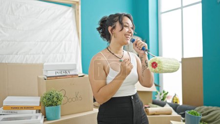 Foto de Joven hermosa mujer hispana sonriendo con confianza cantando canción usando rodillo de pintura como un micrófono en casa nueva - Imagen libre de derechos
