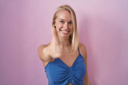 Photo for Young caucasian woman standing over pink background smiling friendly offering handshake as greeting and welcoming. successful business. - Royalty Free Image