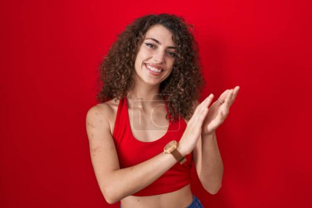 Foto de Mujer hispana con el pelo rizado de pie sobre fondo rojo aplaudiendo y aplaudiendo felices y alegres, sonrientes manos orgullosas juntas - Imagen libre de derechos