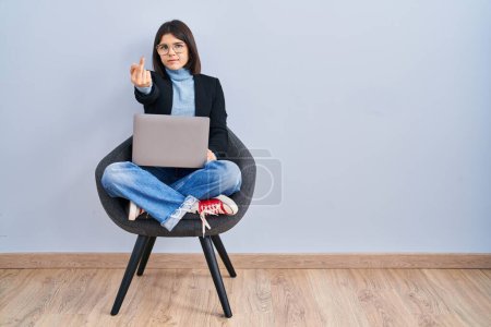 Foto de Young hispanic woman sitting on chair using computer laptop showing middle finger, impolite and rude fuck off expression - Imagen libre de derechos