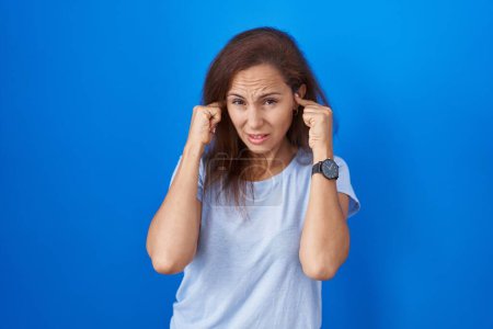 Téléchargez les photos : Femme brune debout sur fond bleu couvrant les oreilles avec des doigts avec une expression agacée pour le bruit de la musique forte. concept sourd. - en image libre de droit