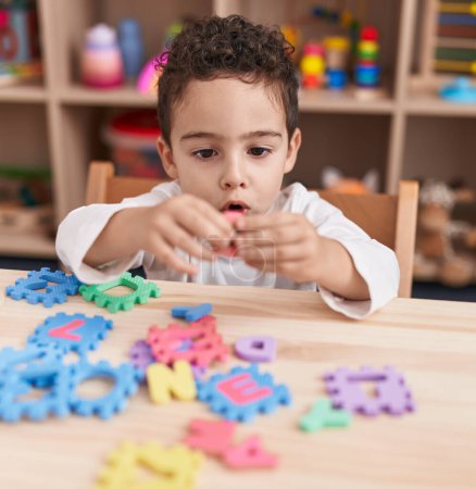 Téléchargez les photos : Adorable garçon hispanique jouant avec le jeu de puzzle de vocabulaire assis sur la table à la maternelle - en image libre de droit