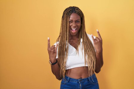 Photo for African american woman with braided hair standing over yellow background shouting with crazy expression doing rock symbol with hands up. music star. heavy music concept. - Royalty Free Image