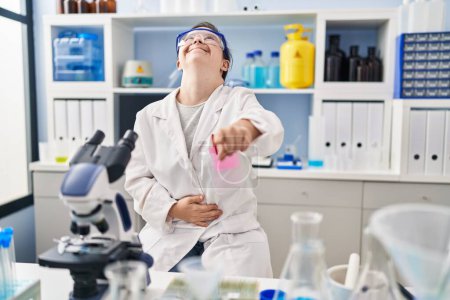 Foto de Chica hispana con síndrome de Down que trabaja en el laboratorio científico riéndose de ti, señalando con el dedo a la cámara con la mano sobre el cuerpo, expresión de vergüenza - Imagen libre de derechos