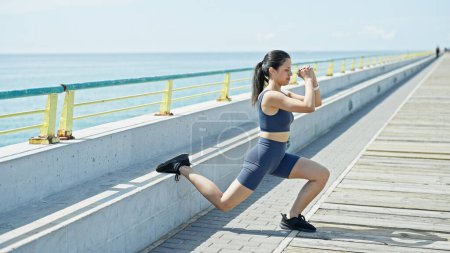 Photo for Young beautiful hispanic woman wearing sportswear training legs exercise at seaside - Royalty Free Image
