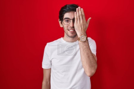 Téléchargez les photos : Young hispanic man standing over red background covering one eye with hand, confident smile on face and surprise emotion. - en image libre de droit