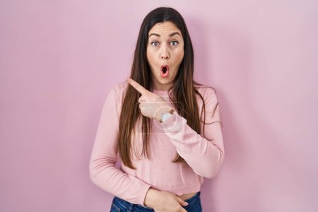Photo for Young brunette woman standing over pink background surprised pointing with finger to the side, open mouth amazed expression. - Royalty Free Image