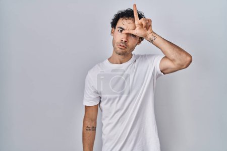 Photo for Hispanic man standing over isolated background making fun of people with fingers on forehead doing loser gesture mocking and insulting. - Royalty Free Image