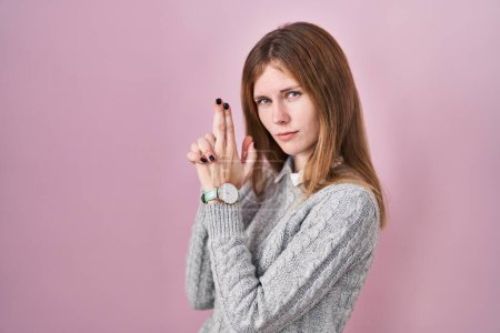 Foto de Hermosa mujer de pie sobre fondo rosa sosteniendo un arma simbólica con gesto de mano, jugando a matar armas de fuego, cara enojada - Imagen libre de derechos