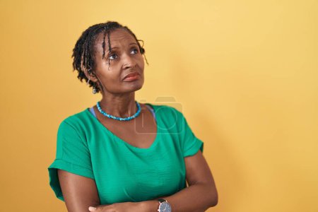 Téléchargez les photos : Femme africaine avec dreadlocks debout sur fond jaune regardant sur le côté avec les bras croisés convaincu et confiant - en image libre de droit