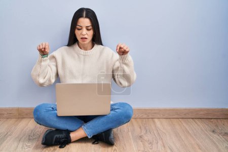 Téléchargez les photos : Jeune femme utilisant ordinateur portable assis sur le sol à la maison pointant vers le bas avec les doigts montrant la publicité, visage surpris et bouche ouverte - en image libre de droit