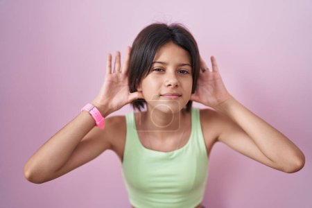 Téléchargez les photos : Jeune fille debout sur fond rose essayant d'entendre les deux mains sur le geste de l'oreille, curieux de commérages. problème auditif, sourd - en image libre de droit