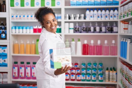 Photo for African american woman pharmacist smiling confident holding vitamin package at pharmacy - Royalty Free Image