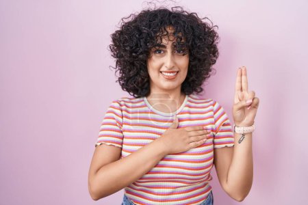 Photo for Young middle east woman standing over pink background smiling swearing with hand on chest and fingers up, making a loyalty promise oath - Royalty Free Image