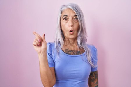 Téléchargez les photos : Femme du moyen âge avec des tatouages debout sur fond rose surpris pointant du doigt sur le côté, bouche ouverte expression étonnée. - en image libre de droit