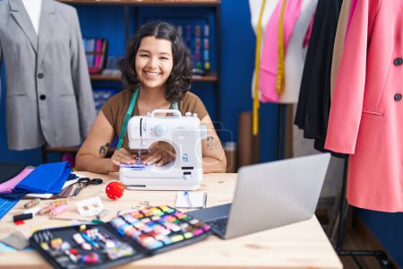 Téléchargez les photos : Jeune femme tailleur utilisant machine à coudre et ordinateur portable au studio de couture - en image libre de droit