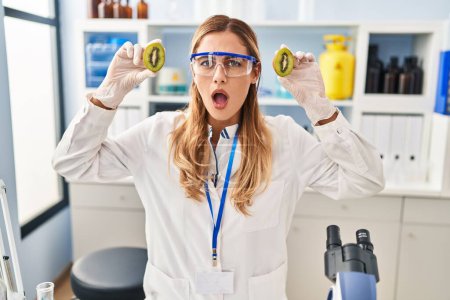 Téléchargez les photos : Jeune scientifique blonde femme travaillant avec de la nourriture au laboratoire en état de choc visage, l'air sceptique et sarcastique, surpris avec la bouche ouverte - en image libre de droit