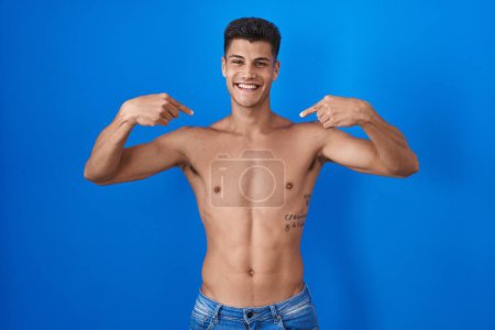 Téléchargez les photos : Jeune homme hispanique debout torse nu sur fond bleu regardant confiant avec le sourire sur le visage, se montrant du doigt fier et heureux. - en image libre de droit
