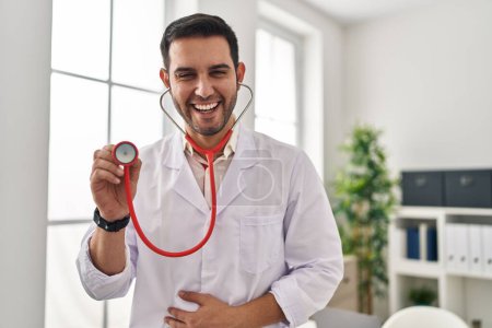 Foto de Joven médico hispano con barba sosteniendo estetoscopio auscultando sonriendo y riendo en voz alta porque gracioso chiste loco. - Imagen libre de derechos
