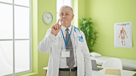 Foto de Médico hombre de pelo gris de mediana edad de pie con expresión seria diciendo no con el dedo en la clínica - Imagen libre de derechos