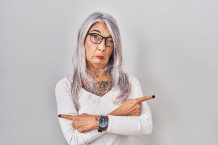 Téléchargez les photos : Femme d'âge moyen avec les cheveux gris debout sur fond blanc pointant vers les deux côtés avec les doigts, direction différente en désaccord - en image libre de droit