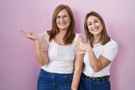 Foto de Madre e hija hispanas vistiendo una camiseta blanca casual sobre fondo rosa asombradas y sonriendo a la cámara mientras se presentan con la mano y señalando con el dedo. - Imagen libre de derechos