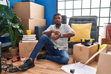 Téléchargez les photos : Homme afro-américain assis sur le sol à la nouvelle maison à la hâte pointant vers regarder le temps, l'impatience, bouleversé et en colère pour retard de délai - en image libre de droit