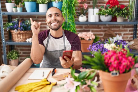 Foto de Hombre hispano con barba trabajando en floristería con smartphone sonriendo feliz señalando con la mano y el dedo hacia un lado - Imagen libre de derechos