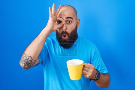 Foto de Joven hombre hispano con barba y tatuajes bebiendo una taza de café haciendo buen gesto conmocionado con la cara sorprendida, los ojos mirando a través de los dedos. expresión incrédula. - Imagen libre de derechos
